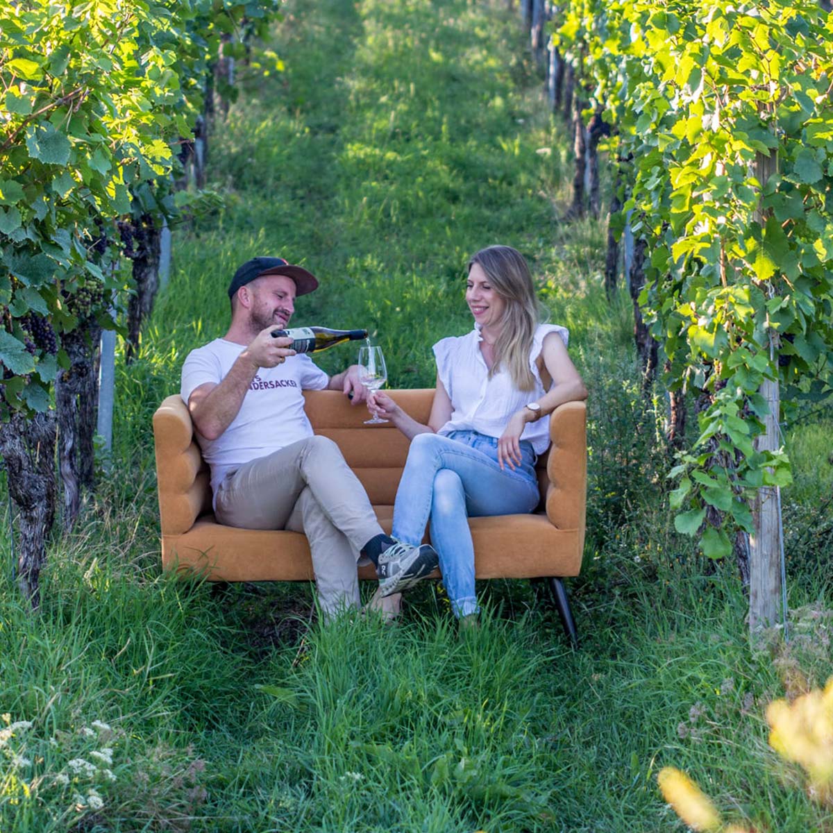Mann und Frau lachen auf einem Sofa im Weinberg, er schenkt Wein ein.